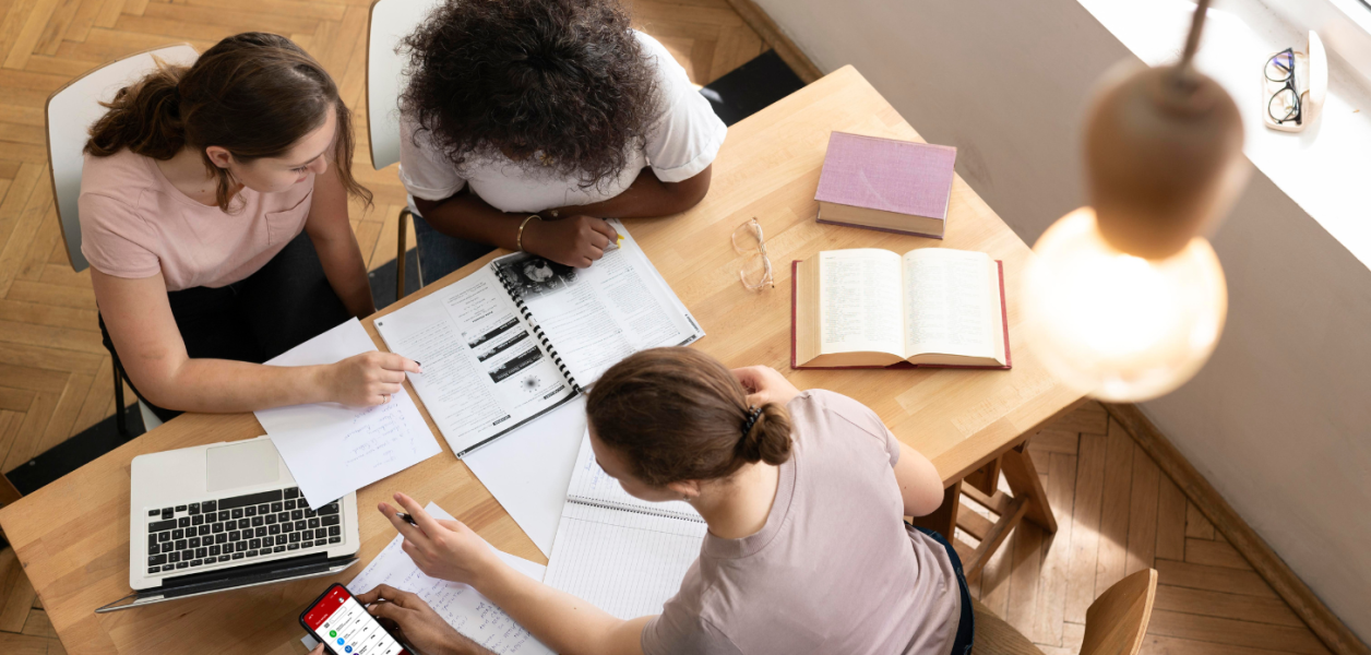 trois filles qui travaillent sur une table sur un projet pour leur étude en BUT infocom. Elle travaillent avec un ordinateur et des carnets